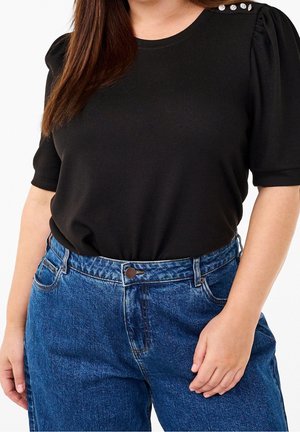Femme portant un haut noir à manches courtes avec des boutons décoratifs sur l'épaule et un jean bleu taille haute, une main posée sur la hanche.