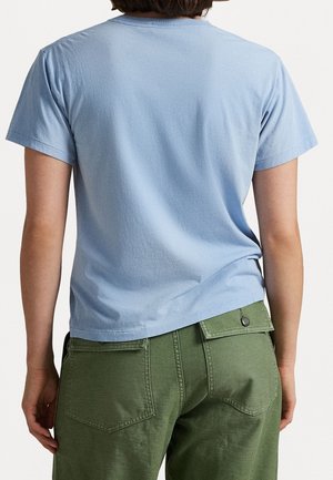 Persoon die een lichtblauw T-shirt met korte mouwen en een groene broek met achterzakken draagt, gezien van achteren tegen een effen achtergrond.