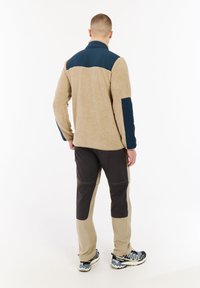 Muž v béžové a námořnické fleecové bundě, dvoubarevných tmavě a světle béžových kalhotách a černo-bílých turistických botách, stojící zády.