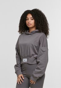 Szara, skrócona bluza z kapturkiem, wykonana z miękkiego materiału, z regulowanym kapturem, szerokimi rękawami i naszywką z przodu. W połączeniu z pasującymi szarymi spodniami.