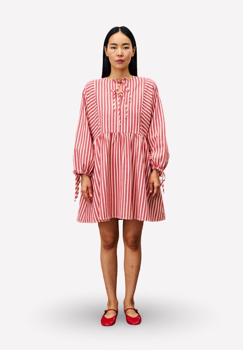 Eine Frau steht und trägt ein rot-weiß gestreiftes Kleid mit langen Ärmeln, Schleifen-Details und roten Ballerinas vor einem schlichten Hintergrund.