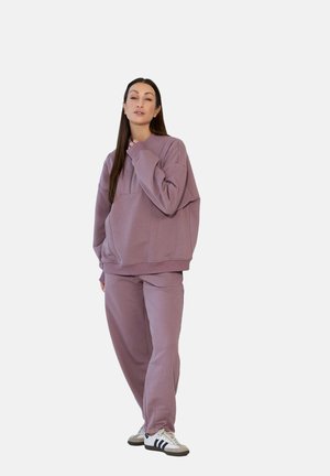 Kobieta ubrana w różowe, oversize'owe bluzy i pasujące spodnie dresowe, nosząca białe i czarne sneakersy, stojąca na tle jednolitego tła.