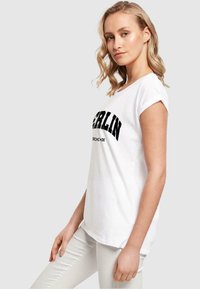 Hvid bomulds t-shirt med en sort "BERLIN" grafik på brystet, korte ærmer og en afslappet pasform. Båret med lyse bukser.