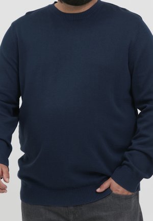 Homme portant un pull à col rond à manches longues bleu marine, une main dans la poche et l'autre détendue le long du corps.