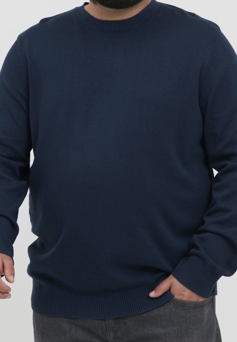 Homme portant un pull à col rond à manches longues bleu marine, une main dans la poche et l'autre détendue le long du corps.