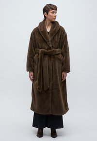 Bruine badjas van imitatiebont met een sjaalkraag, ceintuur en lange mouwen. Gladde textuur, knielang ontwerp, gedragen over een donker outfit en laarzen.