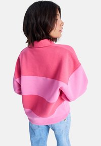 Rosa sweatshirt med fargeblokkdesign i forskjellige nyanser, høy hals og løs passform. Kombinert med lyseblå jeans.