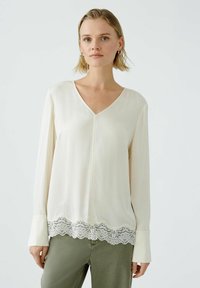 Cremefarbene Bluse mit langen Ärmeln und V-Ausschnitt. Mit einer Spitzenborte am unteren Rand. Glattes Material mit leichtem Glanz und strukturierter Passform.