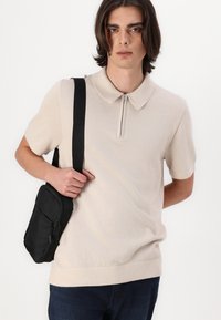 Kremfarget polo skjorte med korte ermer, glidelås fremme, ribbestrikket krage, kombinert med en svart crossoverveske, båret over tettsittende jeans.