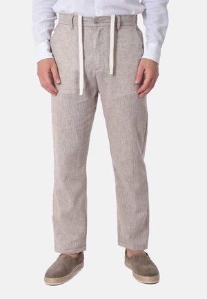 Uomo che indossa pantaloni beige di lino con cordini bianchi, una camicia bianca e scarpe slip-on in camoscio marrone.