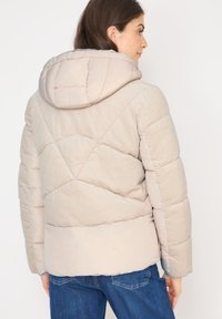 Kvinde iført en beige quiltet jakke med hætte og blå jeans, vist fra ryggen mod en ensfarvet hvid baggrund.