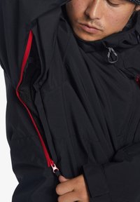 Svart jacka med en röd dragkedja, nätfoder och justerbara dragsko. Texturerad tyg med en strukturerad design och figurnära passform.