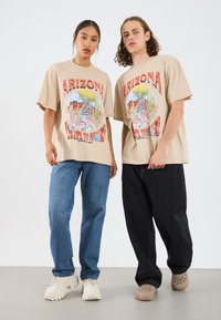 Beige graphic t-shirts featuring a desert scene with a snake, cactus, and text. Model pair wears blue jeans and dark pants with footwear.