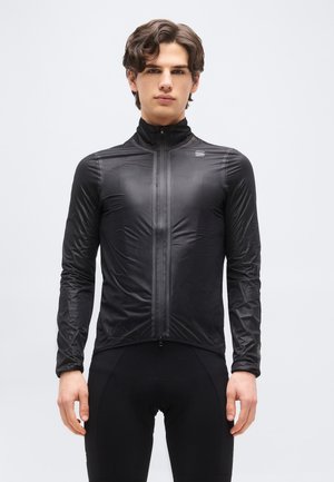 Młody mężczyzna w czarnej, dopasowanej kurtce na suwak i czarnych, obcisłych spodniach, stojący na jasnym, jednolitym tle.