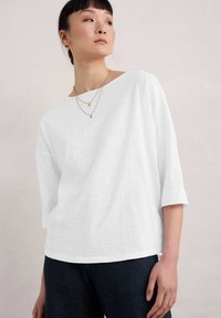 Bílý volný top s širokým výstřihem a tříčtvrtečními rukávy, zkombinovaný s tmavými džíny. Viditelné jsou dvě zlaté náhrdelníky. Hladká textura.