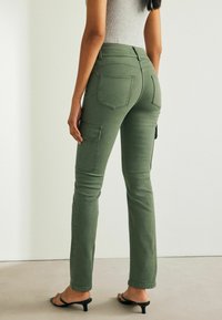 Khaki kalhoty v olivově zeleném denimu, s bočními kapsami, strukturovaným střihem a slim fit nohavicemi, doplněné černými sandály na podpatku.