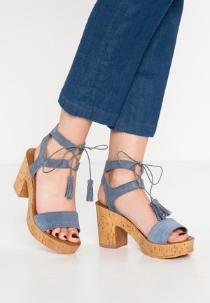 Fødder iført blå ruskindssandaletter med høje hæle, korkplatform og ankelstropper bundet i sløjfer, kombineret med cropped blå denim bukser.