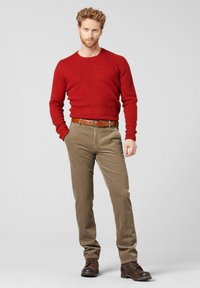 Rode gebreide trui, bruine ribfluwelen broek, bruine leren riem en donkere schoenen. Model staat tegen een neutrale achtergrond, waardoor de outfit wordt benadrukt.