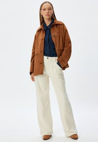 Chaqueta de ante marrón con cierre de botón, combinada con una camisa de mezclilla azul y pantalones de pierna ancha texturizados en color crema. El calzado es marrón.