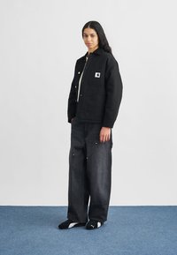 Juoda darbo striukė su priekinėje dalyje esančia kišene, derinama su laisvais tamsiais džinsiniais kelnėmis ir juodais batais. Minimalistinis dizainas su tekstūruotu paviršiumi.