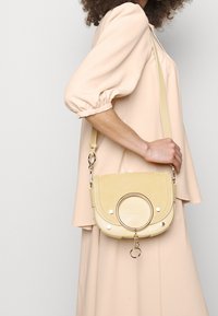 Sac bandoulière jaune avec une forme arrondie, texture lisse, accent de rond en métal et détail en chaîne. La sangle est réglable.