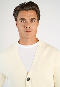 Krämfärgad ribbad cardigan med V-halsdesign, med en enda mörk knappstängning. Bärs över en vit rundhalshimmel.