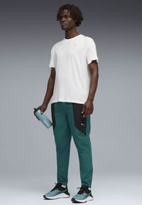 Άνδρας με λευκό t-shirt της Puma, τυρκουάζ και μαύρο παντελόνι φόρμας της Puma, τυρκουάζ και μαύρα παπούτσια τρεξίματος, κρατώντας ένα μπλε μπουκάλι νερού της Puma, στέκεται μπροστά σε γκρίζο φόντο.