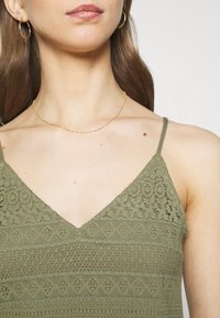Olivově zelený pletený top bez rukávů s texturovaným vzorem, má výstřih do V a tenké ramínka. Nosený jemný zlatý řetízek.