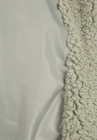 Tejido verde texturizado con un borde de lana esponjosa y con punto, que contrasta con un textil verde claro liso y mate. Se presenta una variación sutil de color.