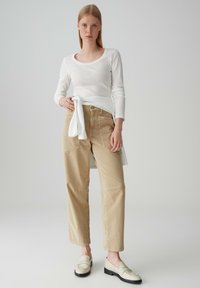 Pantaloni in corduroy beige con una vestibilità ampia, abbinati a una camicia bianca a maniche lunghe legata in vita e mocassini di colore chiaro.