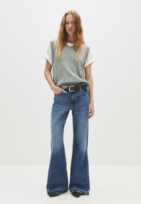 Žena s dlhými červenými vlasmi, oblečená v šedom pletenom svetri cez bielu košeľu s vyhrnutými rukávmi, modrých rozšírených džínsoch, čiernym opaskom a čiernymi topánkami.