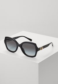Lunettes de soleil noires surdimensionnées avec des verres dégradés, présentant une monture géométrique et un accent en émail doré sur le côté. Fini lisse et brillant.