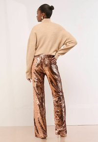 Nő kontyba tűzött hajjal, bézs garbó pulóverben és magas derekú, fényes réz színű flitteres nadrágban áll, kezét a csípőjére téve.