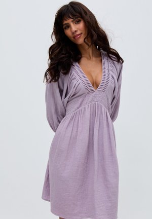 Frau mit langen dunklen Haaren, die ein fliederfarbenes, strukturiertes Langarmkleid mit tiefem V-Ausschnitt und taillierter Passform trägt, posiert vor einem einfarbigen Hintergrund.