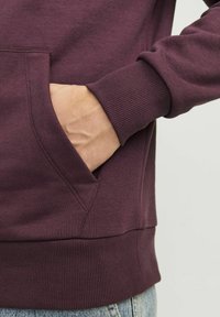 Bordo dukserica s rebrastim manžetama i rubom, s prednjim džepom na struku. Čini se da je materijal mekan s glatkom teksturom.