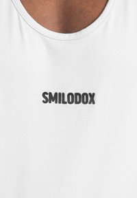 Weißes Baumwoll-Tanktops mit einem runden Ausschnitt und einem auffälligen schwarzen "SMILODOX"-Logo auf der Vorderseite. Glatte Textur, minimalistisches Design.