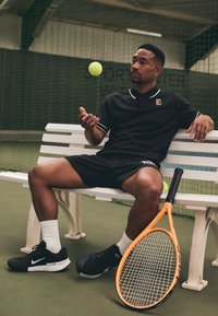 Svart polo-skjorte med hvite detaljer, svarte shorts og svarte Nike-sneakers. Orange tennisracket ved siden av en hvit benk på en innendørs bane.