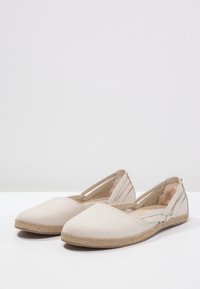 Par de zapatos planos beige de tipo deslizante con puntera cerrada, suelas de textura trenzada y forro interior acolchado, exhibidos sobre una superficie blanca.