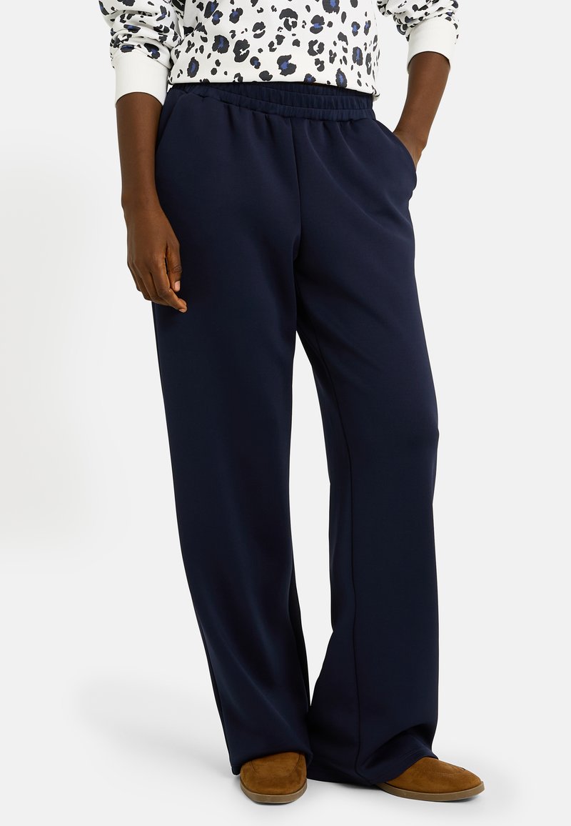 Un pantalon de survêtement bleu marine à la coupe décontractée, avec une taille élastique et des poches latérales, associé à un haut à manches longues à motifs et des chaussures brunes à enfiler.