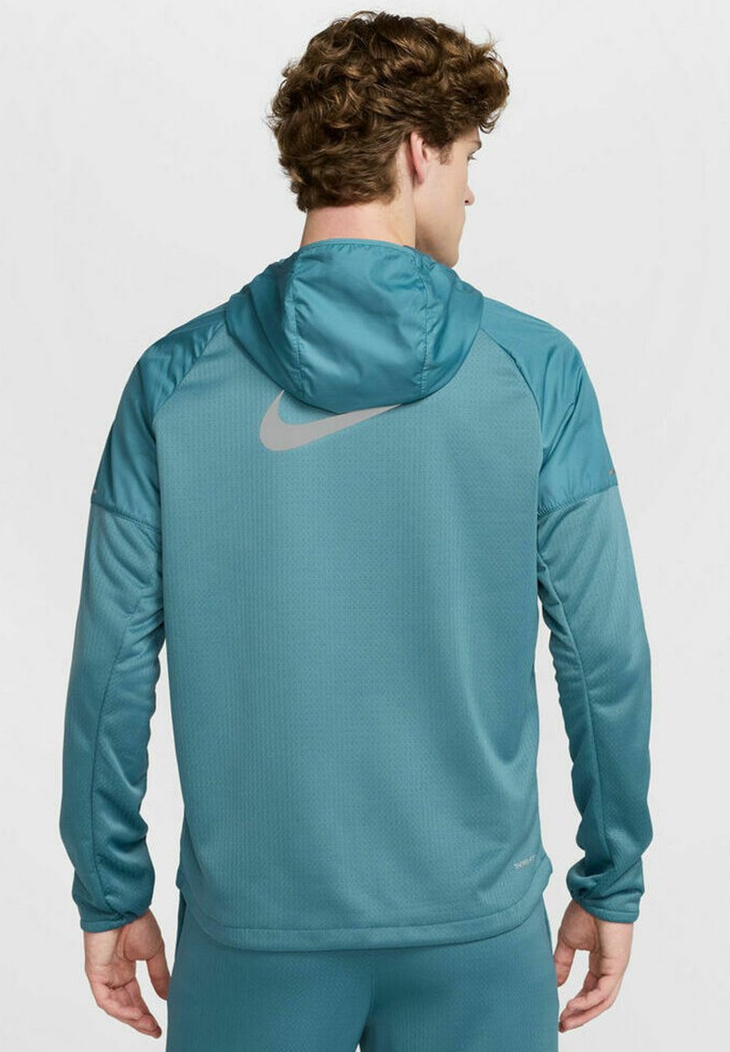 Haut de sport teal avec un tissu texturé, design ajusté et capuche. Présente un logo Nike argenté subtil sur le haut du dos et les manches.