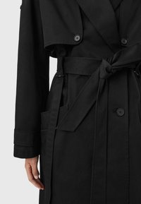 Cappotto nero con grandi bottoni, cintura legata in vita, dettaglio a pattina sul petto, tasca laterale e mano sinistra visibile penzolante accanto al corpo.