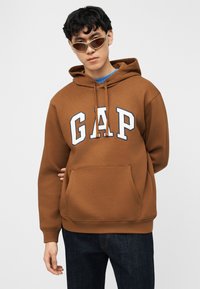 Νέος άνδρας που φοράει καφέ φούτερ GAP, μπλε τζιν και στενά καφέ γυαλιά ηλίου, στέκεται μπροστά σε απλό λευκό φόντο.