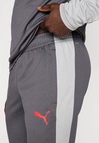 Γκρι παντελόνι φόρμας με αντίθετη ανοιχτό γκρι ρίγα στο πλάι, ελαστική μέση και κόκκινο λογότυπο Puma στον αριστερό μηρό.