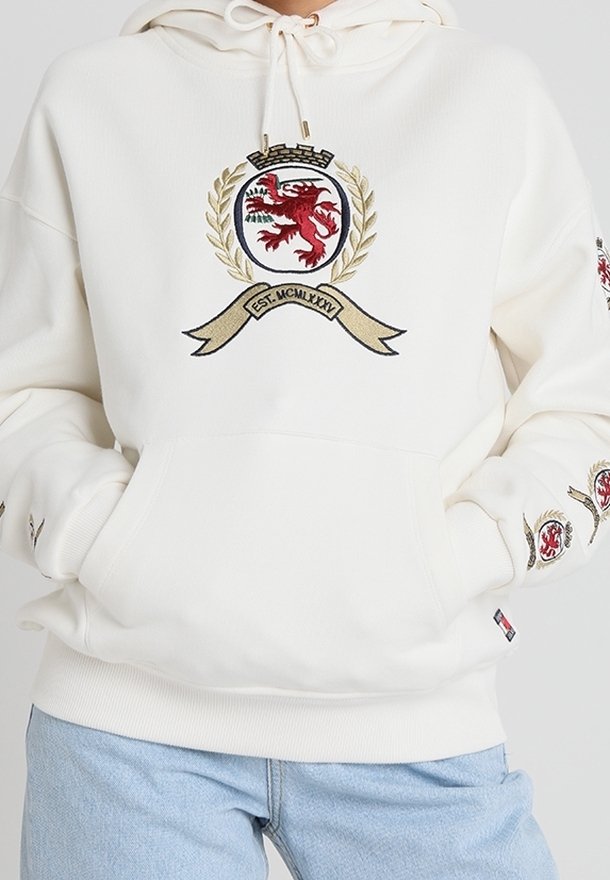 Sudadera color crema con un gran emblema de león rojo y detalles de laurel dorados. Cuenta con capucha ajustable y puños acanalados.