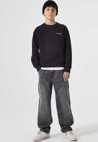 Sudadera morada oscura con cuello redondo y detalle del logotipo en la parte delantera, combinada con jeans de pierna ancha grises y zapatillas blancas. Gorro de punto negro.