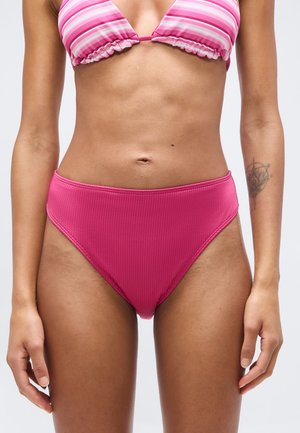 Femeie purtând slip de bikini roz cu dungi verticale, talie înaltă, și sutien de bikini în dungi roz și alb, cu un tatuaj vizibil pe antebrațul stâng.