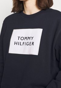 Donna che indossa una felpa scura con una cucitura rettangolare bianca recante il testo "TOMMY HILFIGER" ricamato in lettere nere.