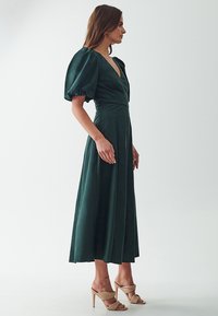 Syvän vihreä midi-mekko, jossa on puffihihat, v-pääntie ja istuva yläosa. Mekossa on virtaava hameosa ja pehmeä pinta, ja se on yhdistetty nuden värisiin korkokenkiin.