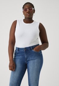 White ribbed tank top paired with dark blue skinny jeans, featuring a pocket and a button closure. Glasses with a pink tint and silver accents.