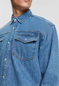 Světle modrá džínová košile s klasickým límečkem, zapínáním na knoflíky, dvěma náprsními kapsami a kontrastním šitím. Texturovaná tkanina.